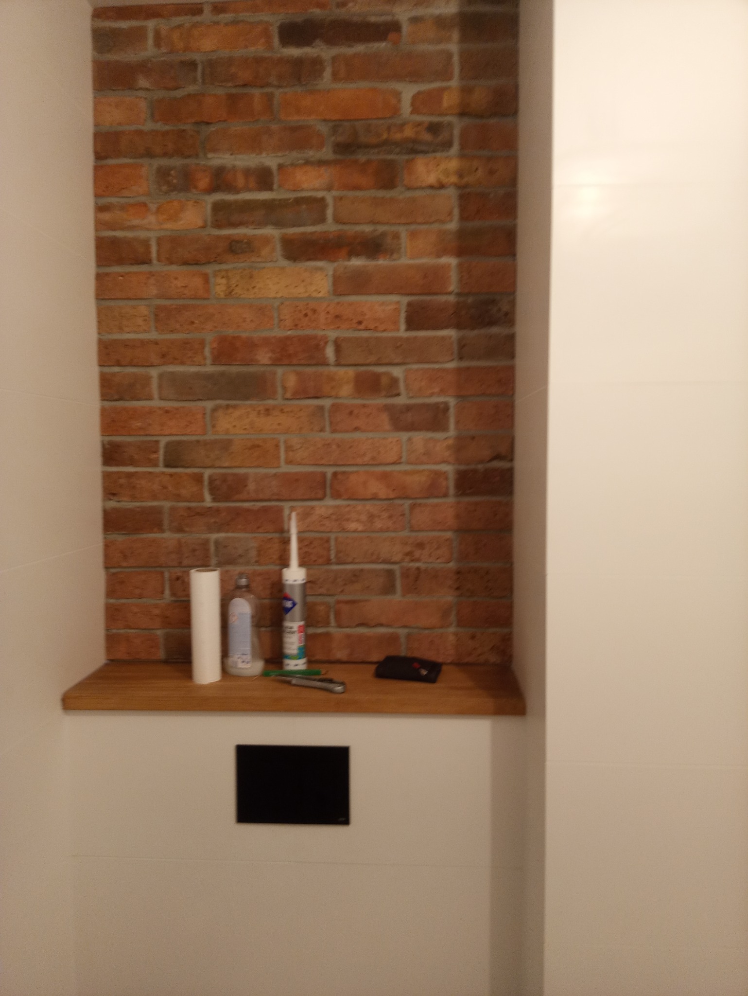 Fragment ściany z dekoracyjną cegłą, drewnianą półką z materiałami budowlanymi i narzędziami, w trakcie prac wykończeniowych w łazience.