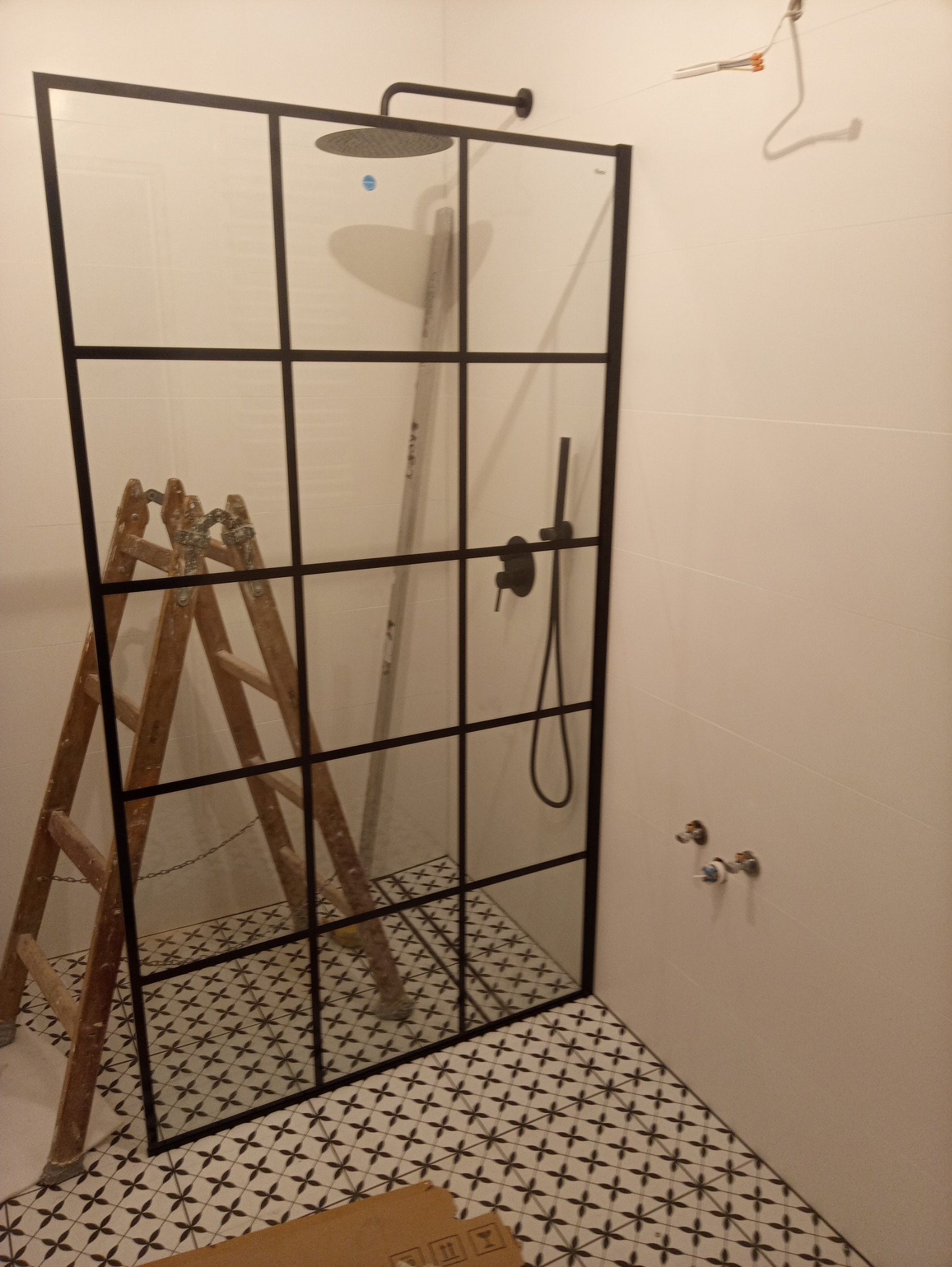 Montaż kabiny prysznicowej typu loft z czarnymi szprosami na podłodze wyłożonej wzorzystymi płytkami, widoczna drabina malarska i elementy instalacji wodnej na ścianie.