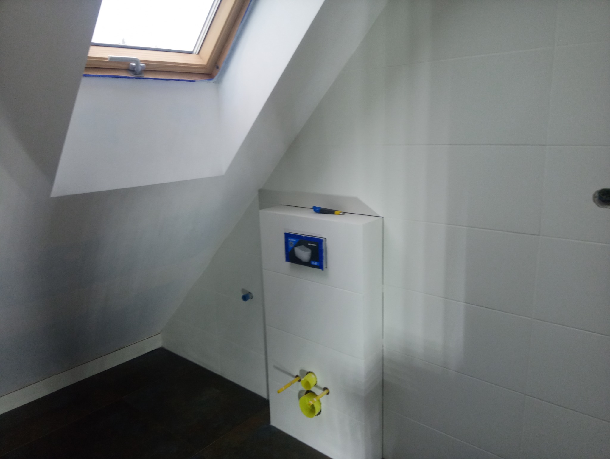 Ukończona łazienka na poddaszu ze skośnym sufitem i oknem dachowym, białe płytki na ścianach, zabudowana toaleta z widocznymi przyłączami wodnymi i narzędziem leżącym na obudowie.