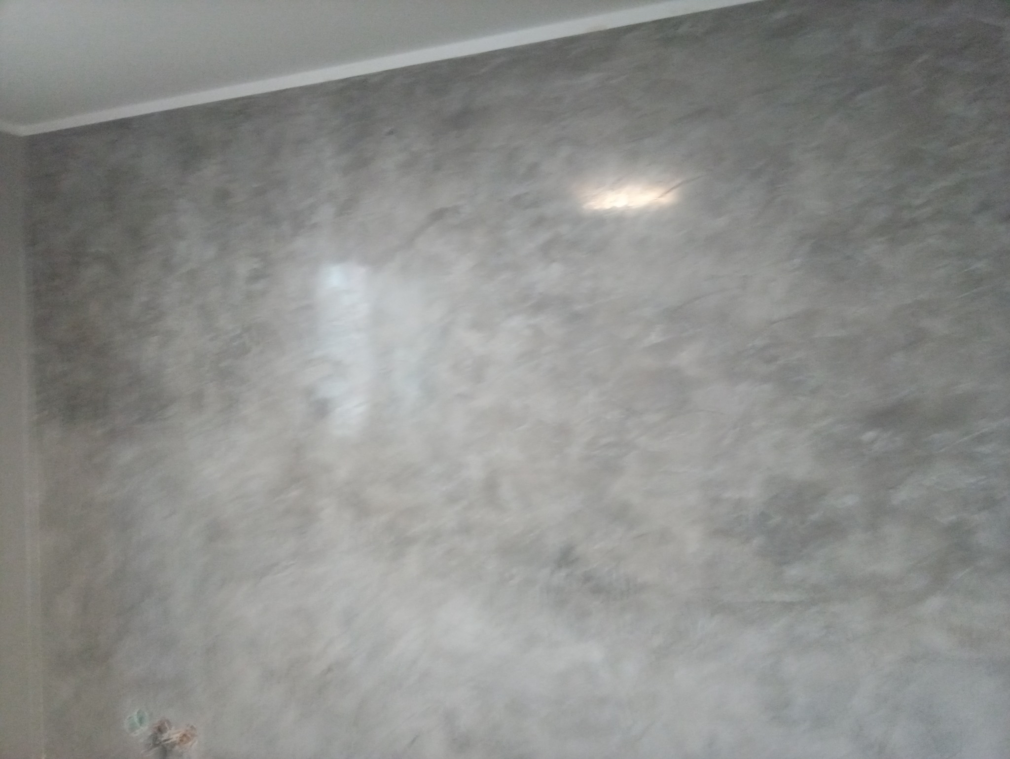 Ściana pokryta szarą, dekoracyjną gładzią z efektem chmur, widoczne refleksy światła.