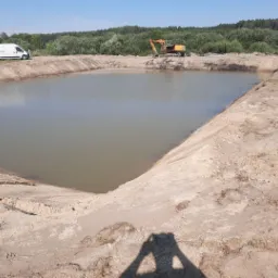 Nowo wykopany staw z żółtą koparką na brzegu i białym busem w tle, widok z góry z cieniem fotografa.