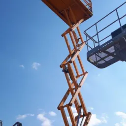 Pomarańczowy podnośnik nożycowy na tle błękitnego nieba z lekkimi obłokami, widok z dołu ukazujący konstrukcję urządzenia i platformę roboczą.