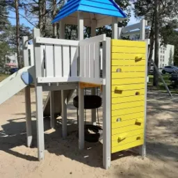 Wielopoziomowy plac zabaw z wieżą krytą niebieskim dachem, ślizgawką i ścianką wspinaczkową w kolorze żółtym, zawieszonymi oponami na łańcuchach, umieszczony na piaszczystym podłożu w otoczeniu drzew.