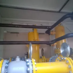 Instalacja gazowa z żółtymi rurami, szarymi elementami i czarnymi rurami zgrzewanymi, umieszczona wewnątrz pomieszczenia z widocznymi ścianami i sufitem.