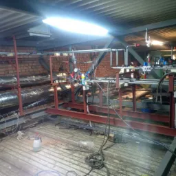 Instalacja rurociągów przemysłowych z elementami pomiarowymi i kontrolnymi, zamontowana na konstrukcji wsporczej z profili stalowych w hali z ceglanymi ścianami i metalowym stropem.