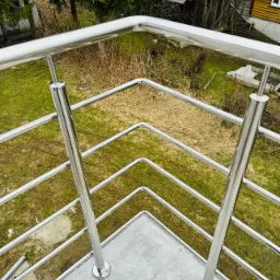 Nowoczesna, błyszcząca balustrada ze stali nierdzewnej na balkonie z szarą płytką, widok z góry na trawnik i zabudowania gospodarcze w tle.