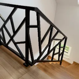 Nowoczesna, geometryczna balustrada schodowa w kolorze czarnym, wykonana w Perle. Widok z góry na drewniane schody i fragment okna.