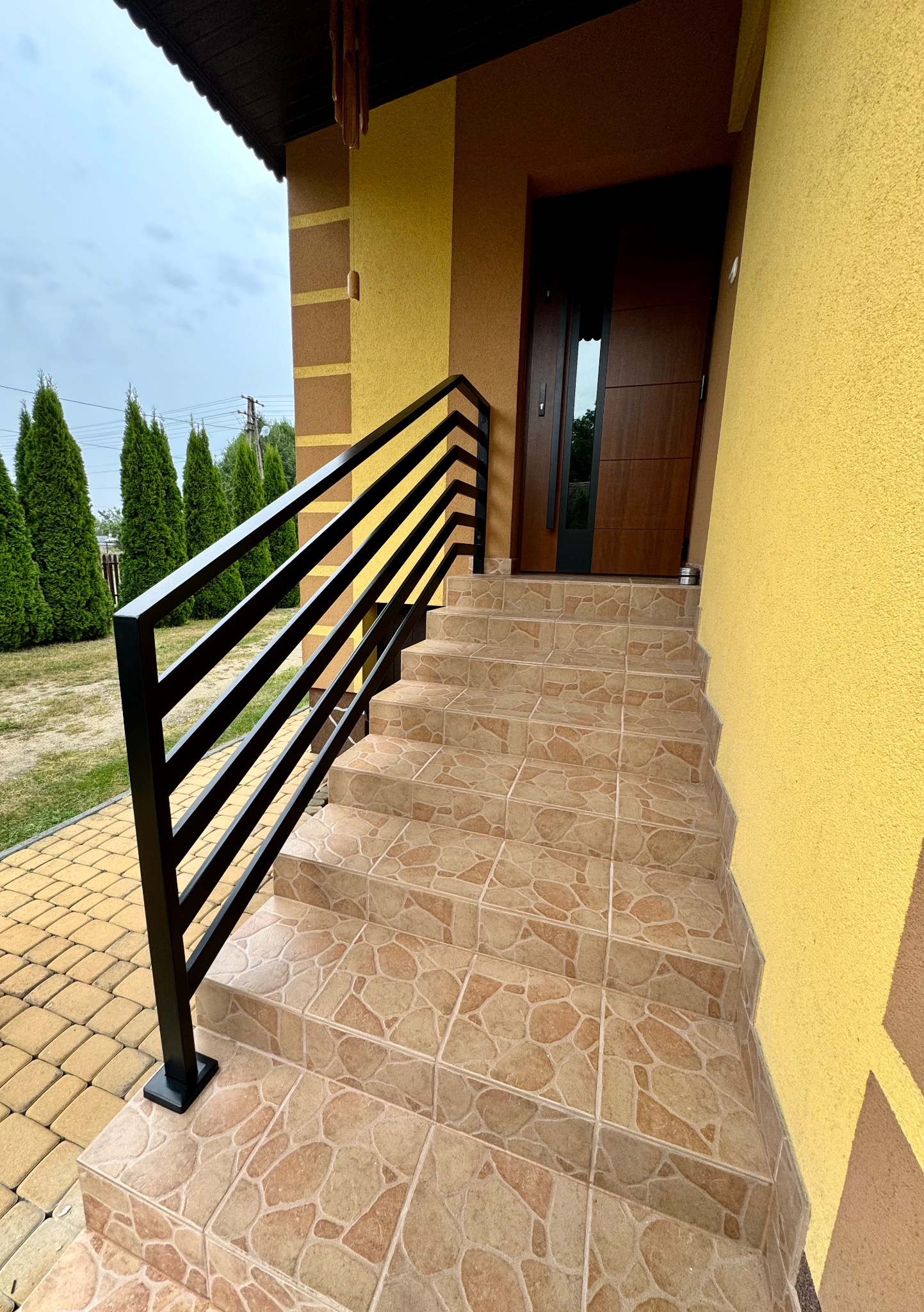 Zewnętrzne schody wejściowe wyłożone płytkami z czarną, metalową barierką z poziomych profili, na tle żółtej elewacji budynku i tujowego żywopłotu.