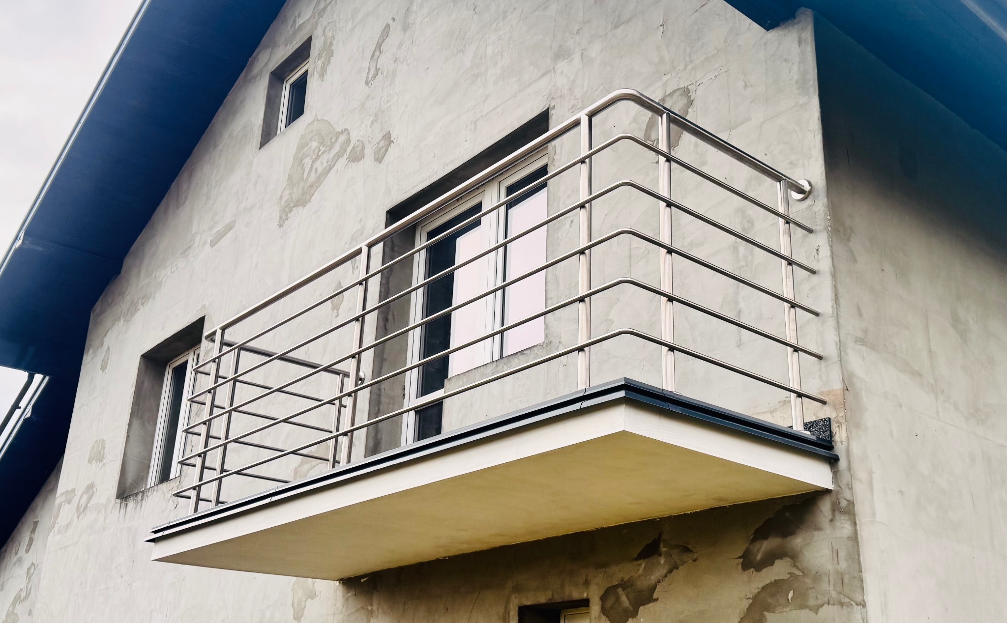 Nowoczesna, metalowa balustrada balkonowa z poziomymi prętami na tle surowej, szarej elewacji budynku. Widok z dołu podkreśla minimalistyczny design i solidne wykonanie.