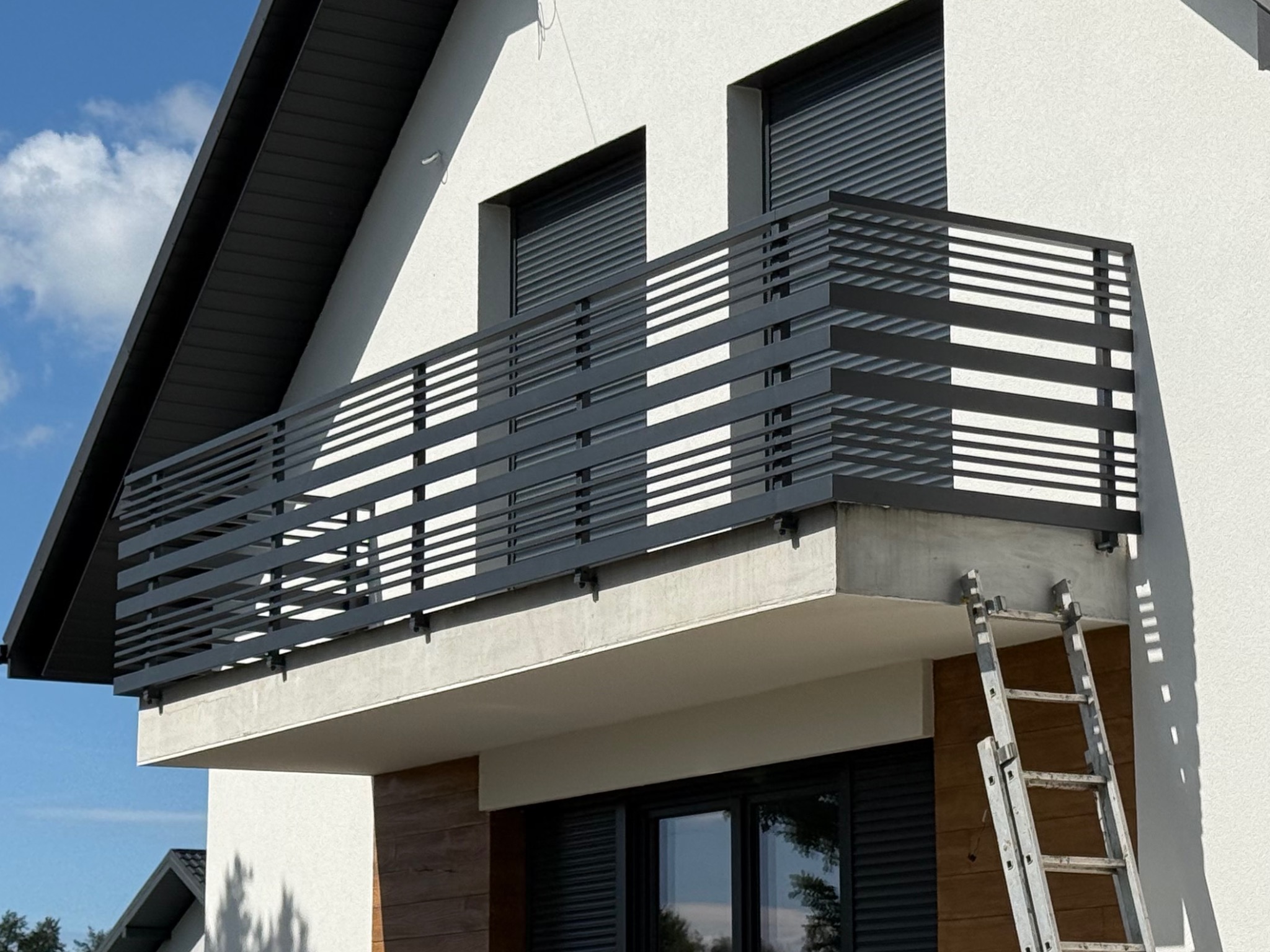 Nowoczesna, pozioma balustrada balkonowa w kolorze antracytowym. Minimalistyczny design z poziomych profili na tle białej elewacji domu.
