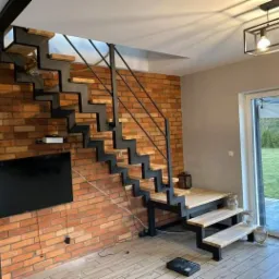 Nowoczesne schody metalowe z drewnianymi stopniami na tle ściany z czerwonej cegły w jasnym wnętrzu domu.