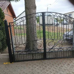 Czarna, metalowa brama wjazdowa z ozdobnymi elementami, widoczna z bliska na tle drzewa, samochodu i budynku.