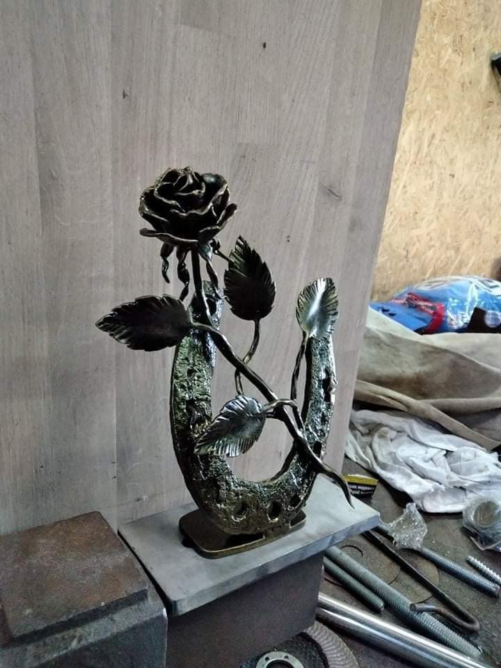 Kuta metalowa róża wyrastająca z podkowy na tle warsztatu, widoczne narzędzia i elementy obróbki metalu.