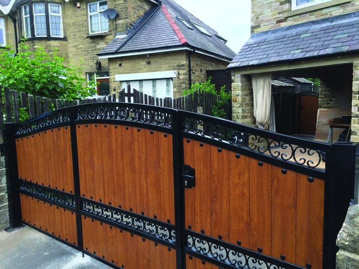 Dwuskrzydłowa brama wjazdowa z drewnianymi panelami i ozdobnymi elementami kutymi, zamontowana przy kamiennym ogrodzeniu, na tle domu z jasnej cegły.