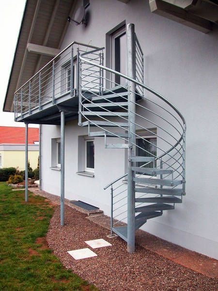 Zewnętrzne metalowe schody spiralne prowadzące na balkon przy białym domu, z widocznym trawnikiem i żwirowym podłożem.