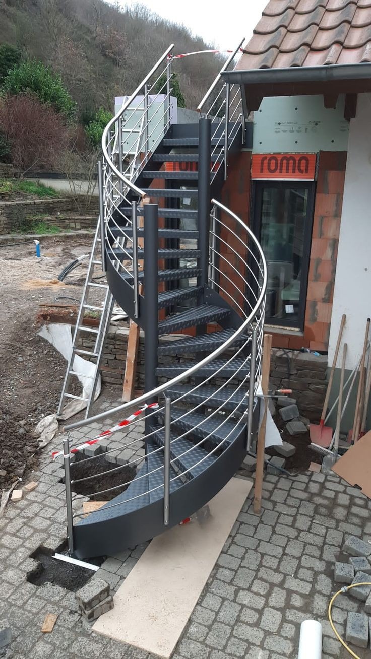 Zewnętrzne, spiralne schody metalowe z poręczą ze stali nierdzewnej, zamontowane przy budynku w trakcie budowy, widoczne narzędzia i materiały budowlane.
