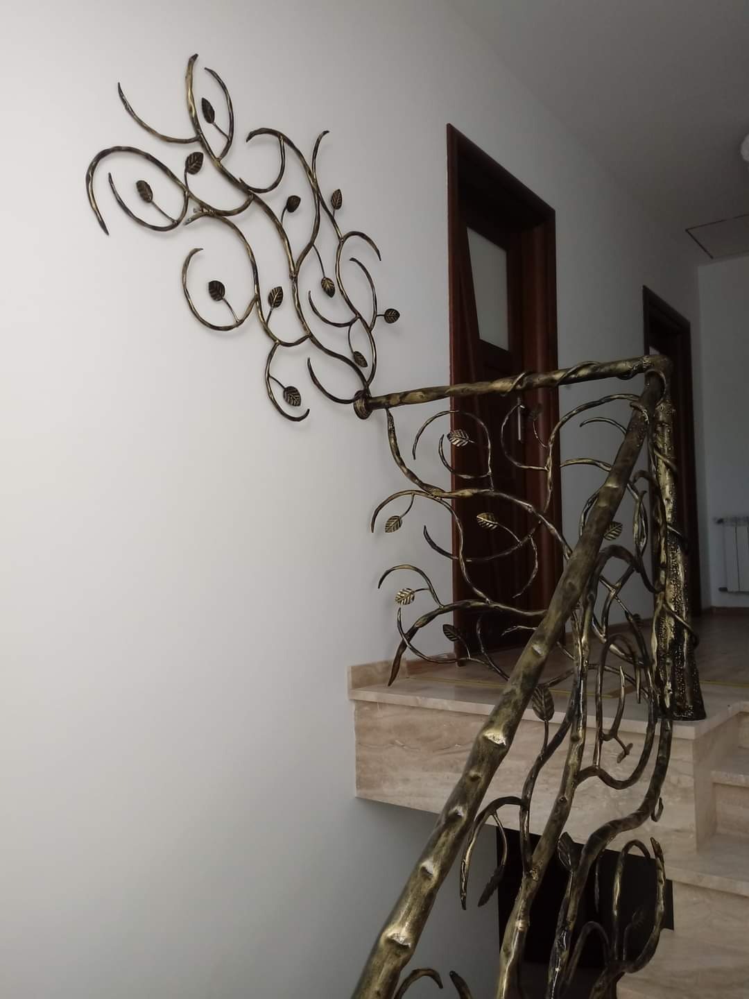 Kuta balustrada schodowa z motywem roślinnym w kolorze starego złota, z dopasowaną dekoracją ścienną, na tle marmurowych stopni i białych ścian.