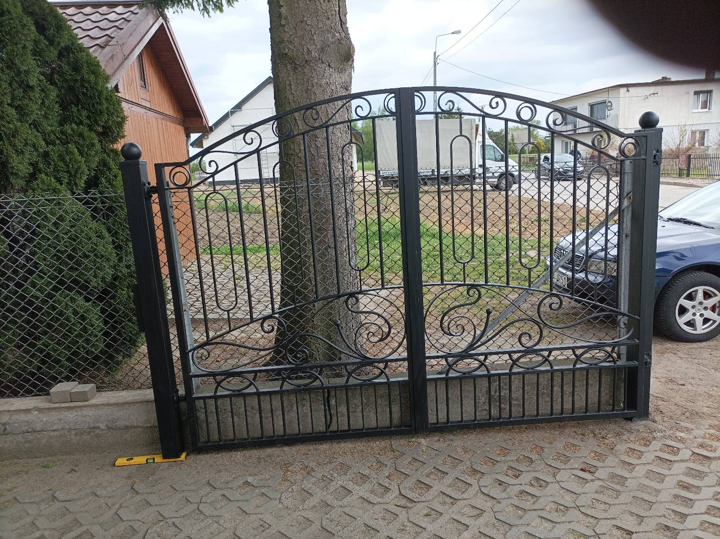 Czarna, metalowa brama wjazdowa z ozdobnymi elementami, widoczna z bliska na tle drzewa, samochodu i budynku.