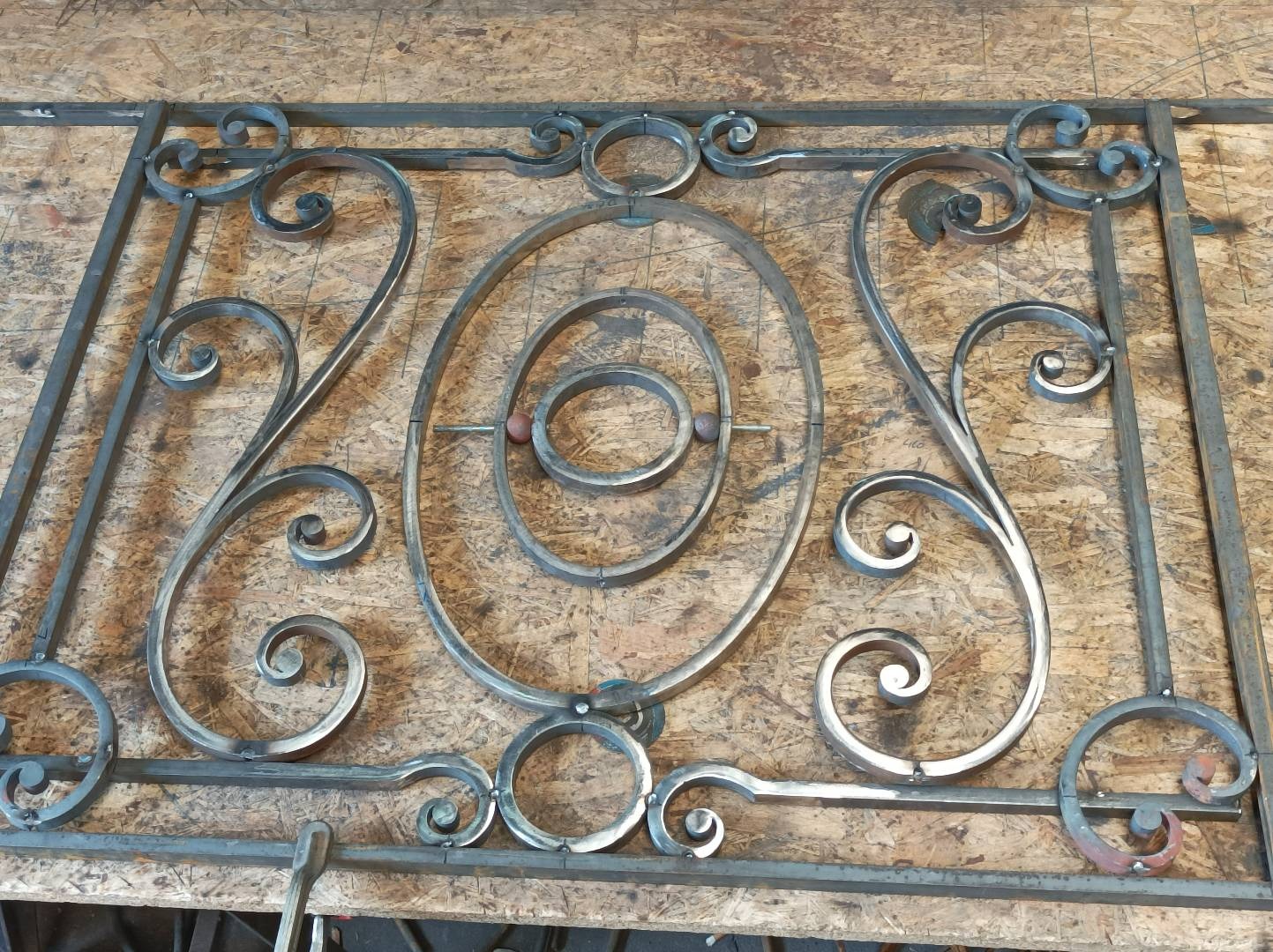 Surowy metalowy element dekoracyjny z ornamentami, prawdopodobnie fragment bramy lub ogrodzenia, leżący na płycie OSB.