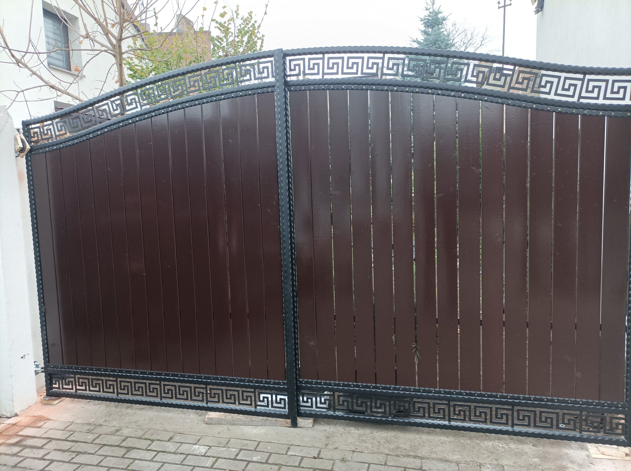 Dwuskrzydłowa brama wjazdowa z brązowych paneli pionowych i czarną metalową ramą z motywem greckim, widoczna z zewnątrz posesji.