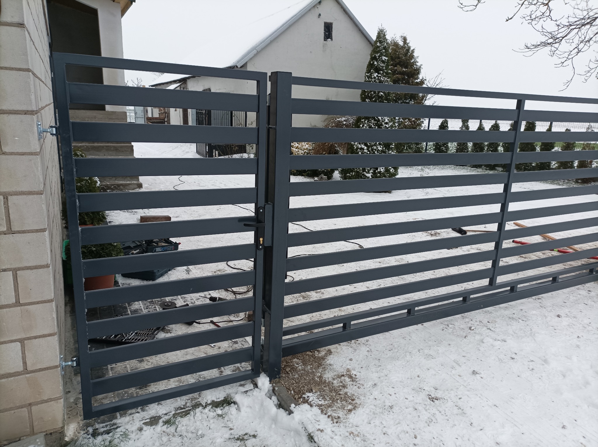 Nowoczesna, szara brama wjazdowa z poziomych profili, częściowo otwarta, widoczna na tle ośnieżonego podwórza i domu. Na pierwszym planie narzędzia i akcesoria montażowe.