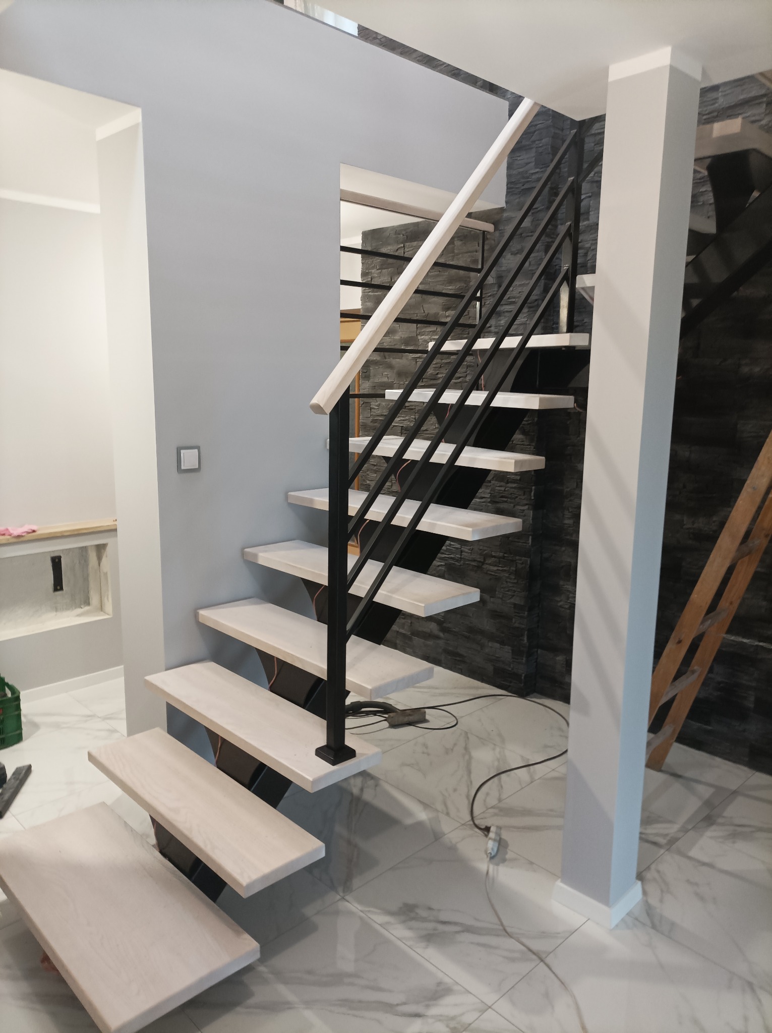 Nowoczesne schody z drewnianymi stopniami i czarną metalową balustradą w jasnym wnętrzu z marmurową podłogą, widoczne kable i drabina w tle.