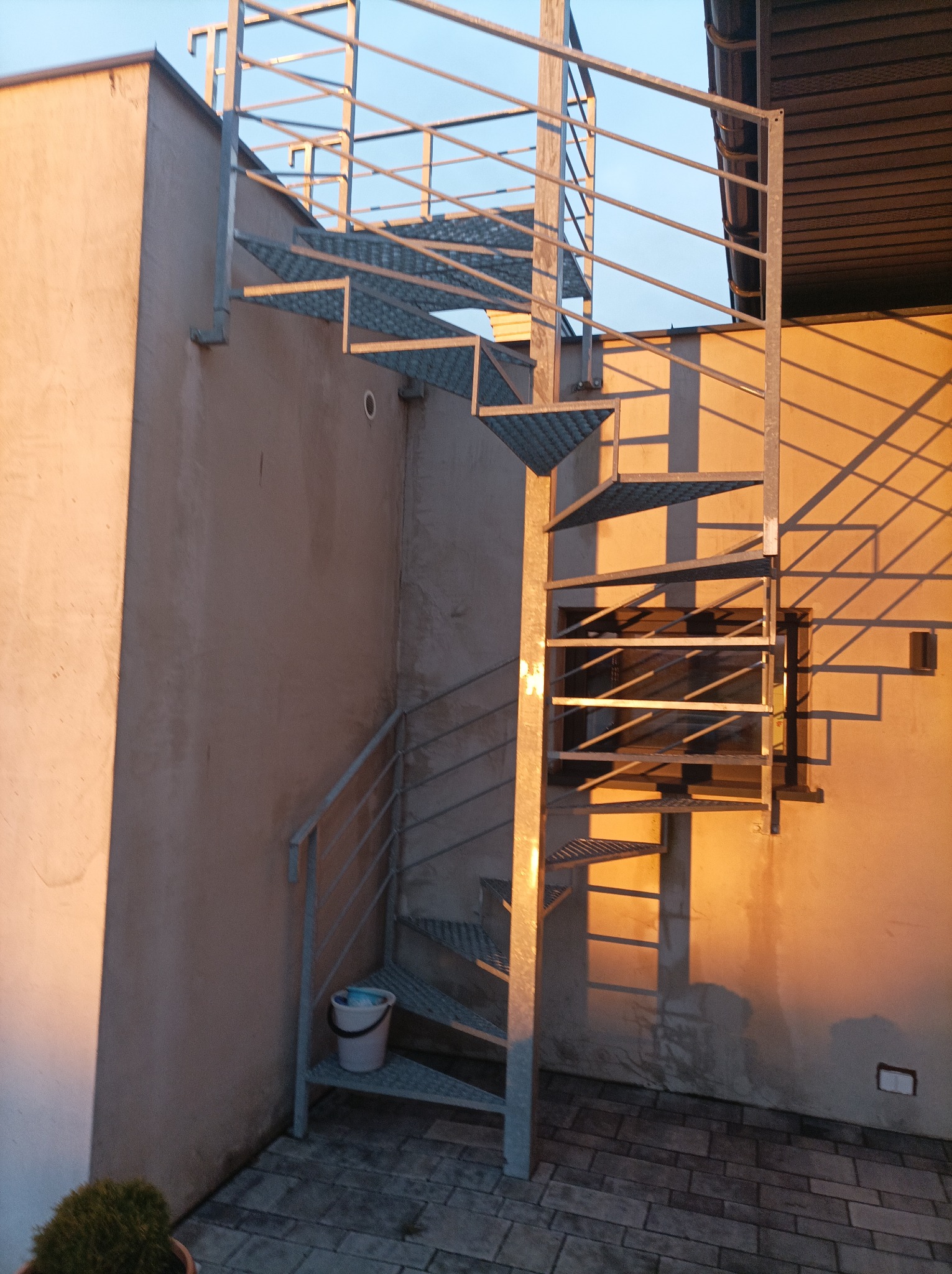 Zewnętrzne metalowe schody spiralne z kratowanymi stopniami, prowadzące na balkon, z wiadrem na dolnym biegu, oświetlone popołudniowym słońcem rzucającym cienie balustrady na ścianę budynku.