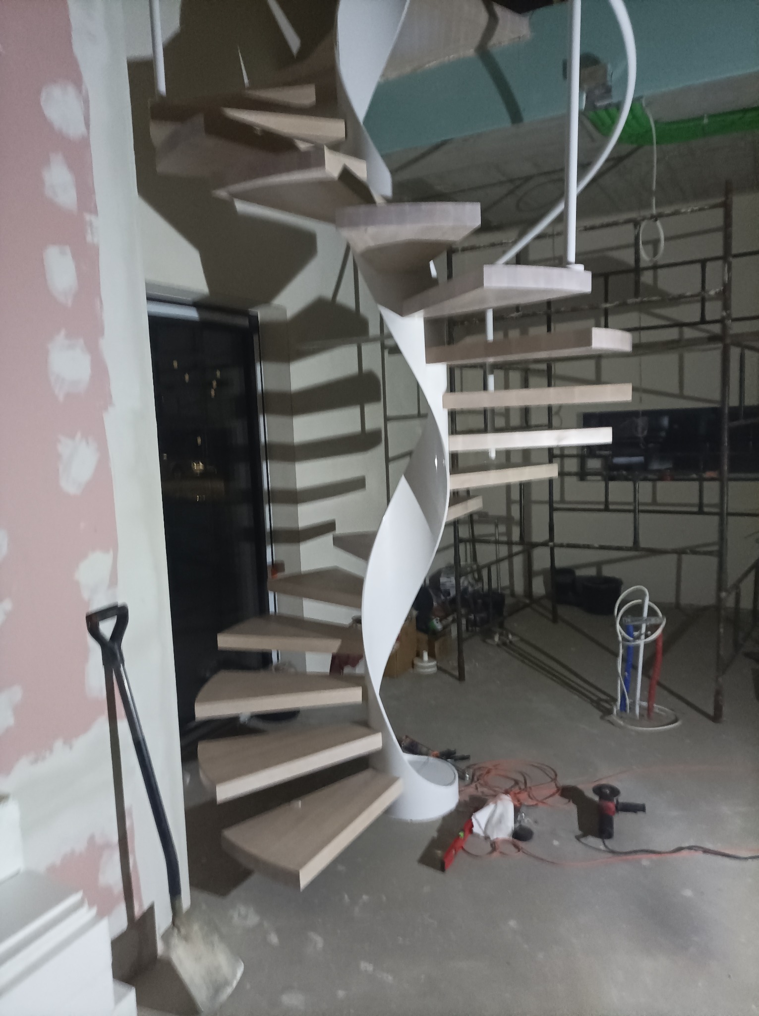 Wewnętrzne schody spiralne z drewnianymi stopniami i białą, metalową konstrukcją nośną w trakcie montażu, widoczne narzędzia i rusztowanie w tle.