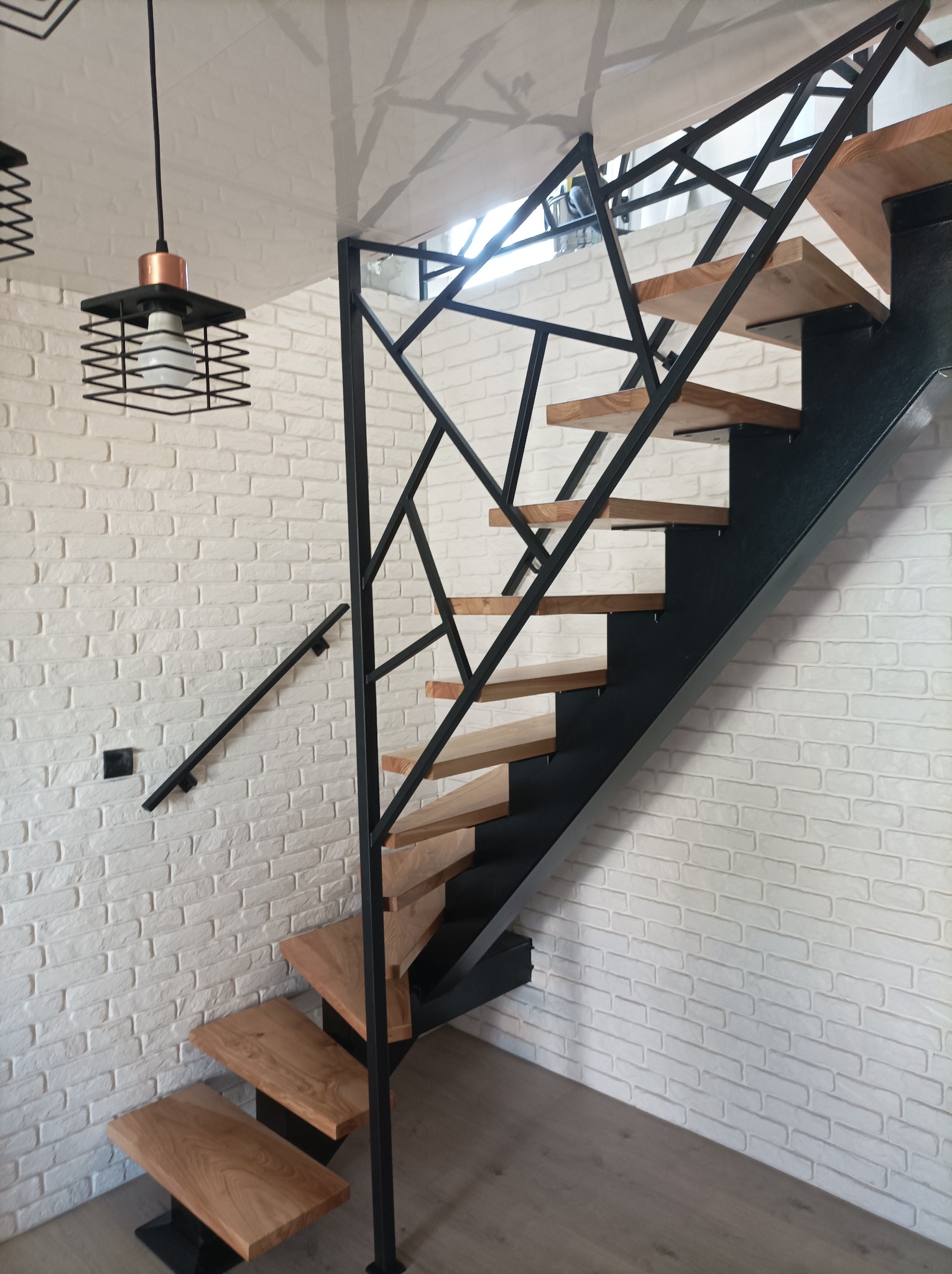 Nowoczesne schody z drewnianymi stopniami i czarną, geometryczną konstrukcją metalową, zamontowane przy ścianie z białej cegły.