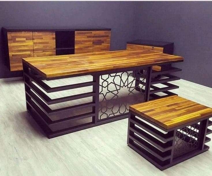 Nowoczesny stół z blatem z drewna klejonego i metalową podstawą z geometrycznymi wycięciami, pasujący do komody i szafki RTV w tle, tworzący spójny zestaw mebli.