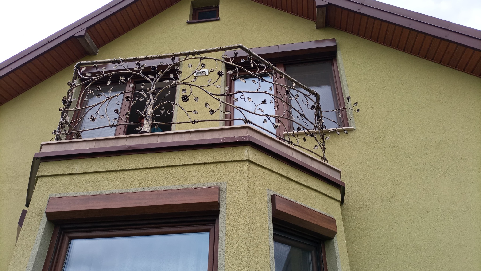 Kuty balkon z dekoracyjną balustradą w kształcie drzewa z różami, zamontowany na budynku o oliwkowej elewacji i brązowej stolarce okiennej.