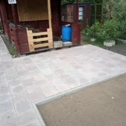 Nowo ułożona, jasnoszara nawierzchnia z kostki brukowej przed drewnianą altaną z niebieską beczką w tle.