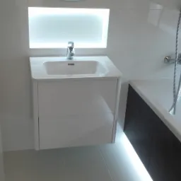 Nowoczesna łazienka z podświetlaną umywalką na szafce, białą wanną z ciemnymi płytkami i chromowaną baterią natryskową na tle białych płytek.