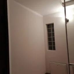 Świeżo pomalowane ściany na gładko w łazience z oknem z luksferów, widoczna kratka wentylacyjna i brązowe płytki podłogowe.