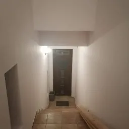 Klatka schodowa z brązowymi płytkami i świeżo pomalowanymi białymi ścianami, widoczny fragment drewnianej listwy przypodłogowej i drzwi wejściowe.