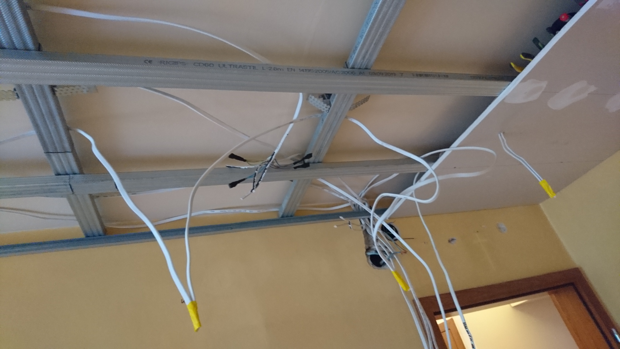 Widok z dołu na sufit podwieszany w trakcie montażu: metalowa konstrukcja, białe kable elektryczne z żółtymi końcówkami, częściowo zamontowana płyta gipsowo-kartonowa.