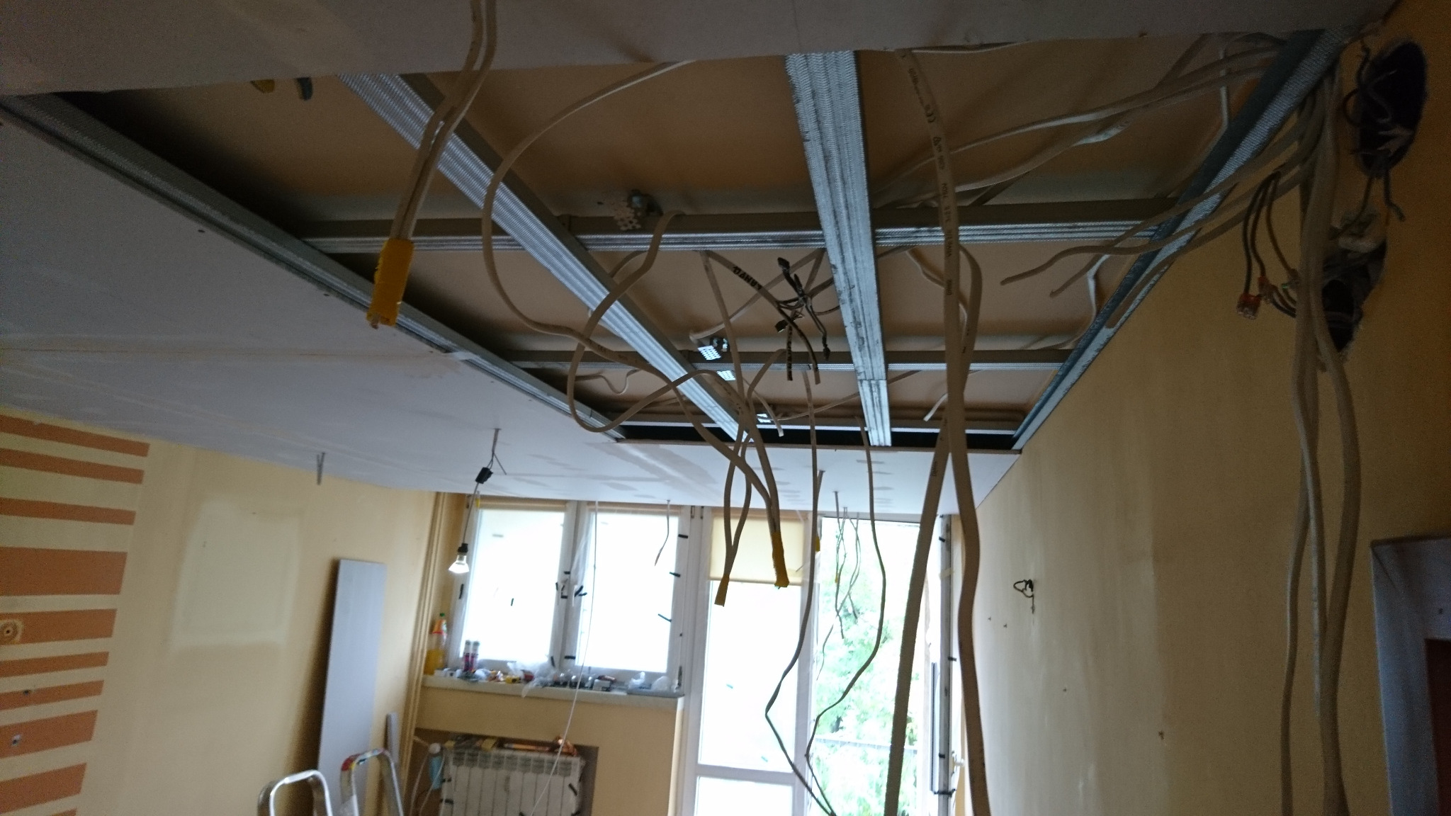 Sufit podwieszany w trakcie montażu z widocznymi przewodami elektrycznymi, metalową konstrukcją i częściowo zamontowanymi płytami gipsowymi. Widoczne okno w tle.