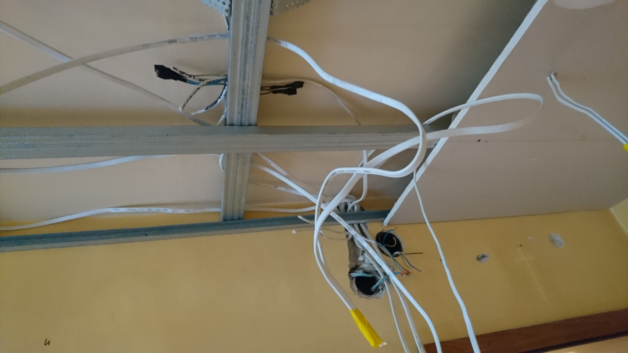 Widok z dołu na montaż sufitu podwieszanego z widocznymi kablami elektrycznymi wystającymi z otworów w suficie, żółte osłonki na końcach kabli, metalowa konstrukcja sufitu.