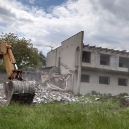 EKVADOR Mateusz Osiński - Żółta koparka z operatorem w kabinie, kruszy betonowy budynek w trakcie rozbiórki, w tle widoczny żuraw budowlany i zachmurzone niebo.