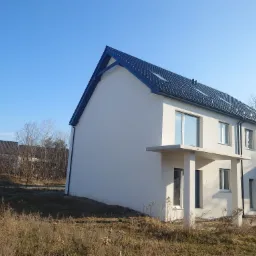 Dwukondygnacyjny, nowo wybudowany bliźniak z ciemną dachówką, jasną elewacją i minimalistycznymi balkonami, sfotografowany w słoneczny dzień na tle błękitnego nieba i jesiennego krajobrazu.