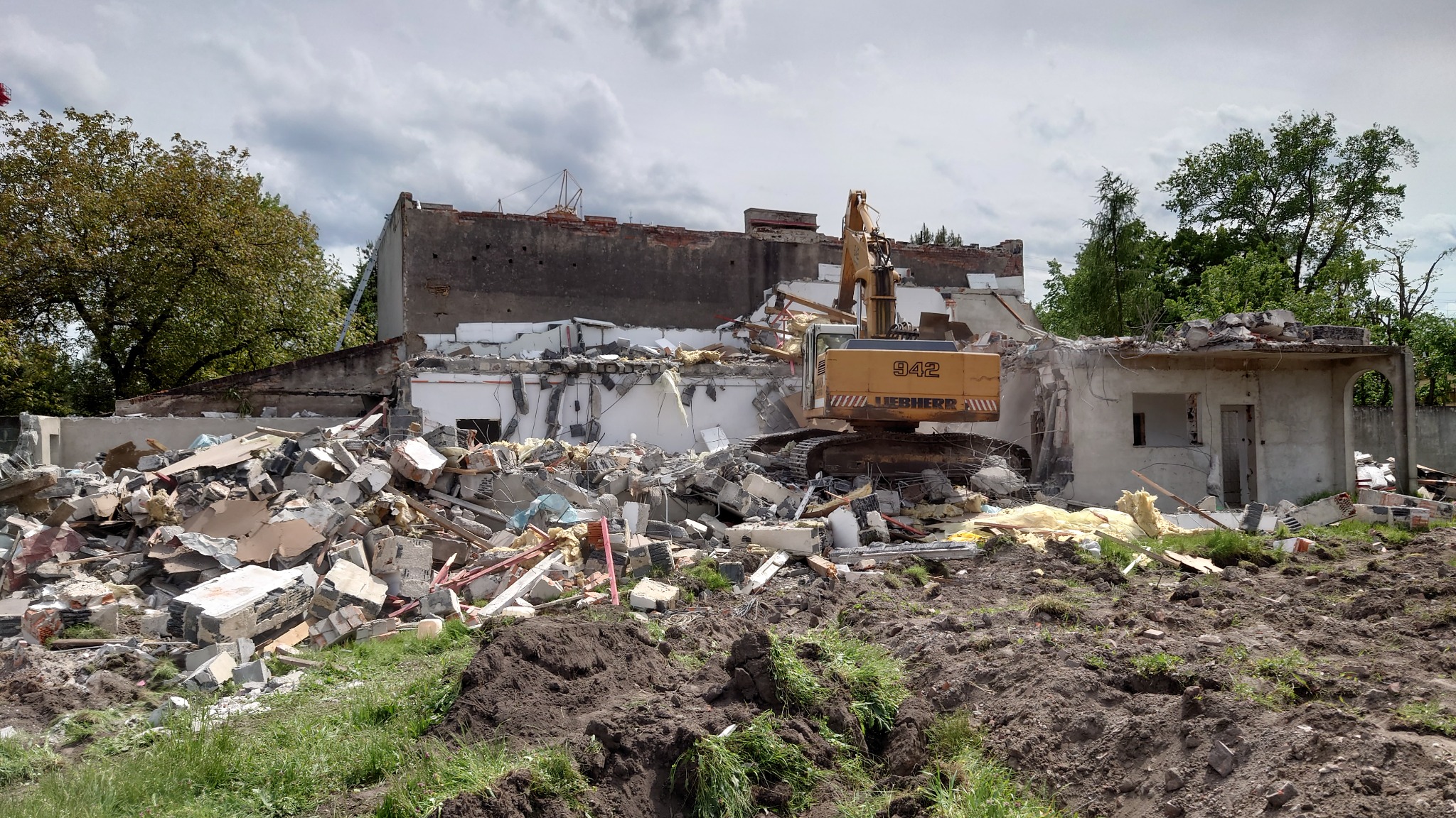 Żółta koparka Liebherr 942 na tle gruzów i częściowo zburzonego budynku z elementami ceglanymi, widoczne drzewa i pochmurne niebo.