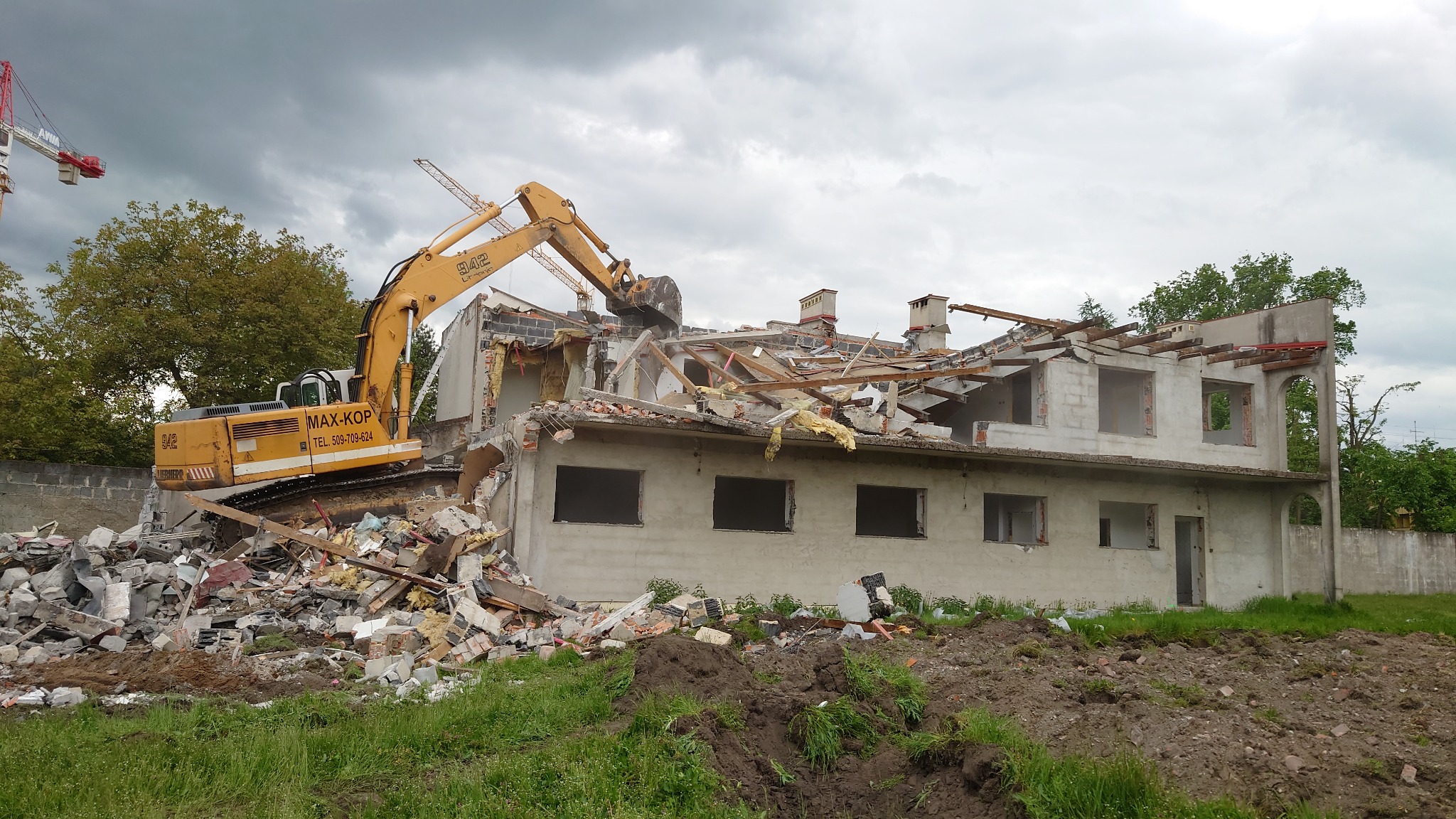 Żółta koparka gąsienicowa MAX-KOP z operatorem demontuje betonowy budynek z widocznymi otworami okiennymi i resztkami dachu, gruzowisko na pierwszym planie, pochmurne niebo w tle.