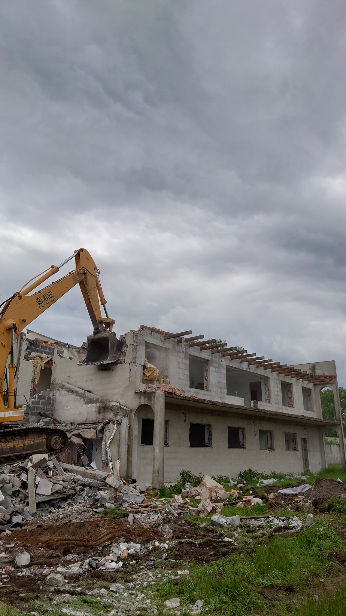Żółta koparka z łyżką na wysokości drugiego piętra rozbiera murowany budynek z odsłoniętymi belkami stropowymi, gruzowisko u podnóża budynku, pochmurne niebo.