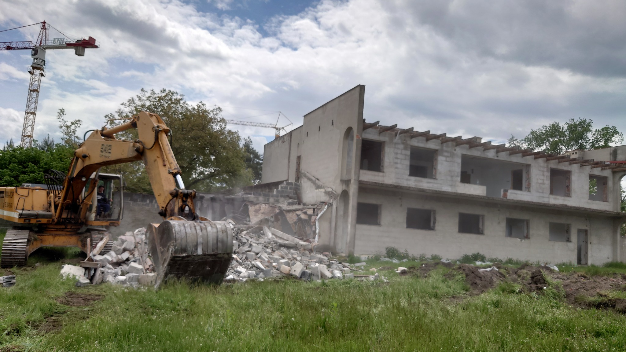 Żółta koparka z operatorem w kabinie, kruszy betonowy budynek w trakcie rozbiórki, w tle widoczny żuraw budowlany i zachmurzone niebo.