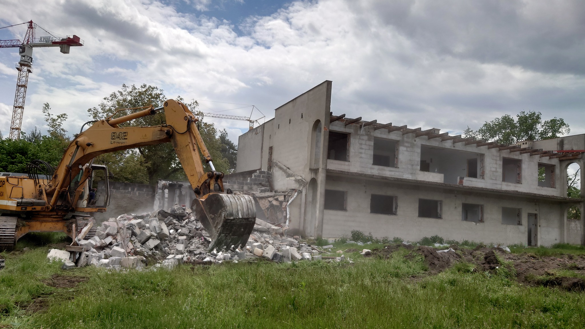 Żółta koparka z łyżką kruszy fragmenty muru budynku w trakcie rozbiórki, w tle widoczny żuraw budowlany i fragmenty konstrukcji budynku.