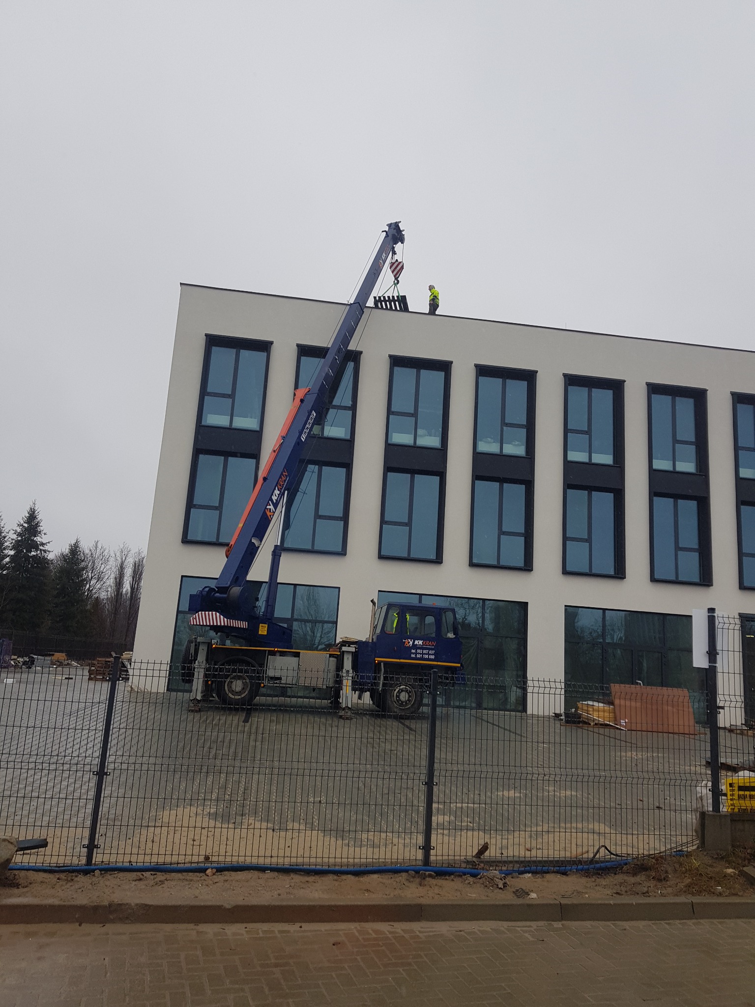 Mobilny dźwig z wysięgnikiem podnosi materiały na dach nowoczesnego budynku z dużymi oknami. Pracownik w żółtym ubraniu roboczym na dachu budynku. Widok zza ogrodzenia.