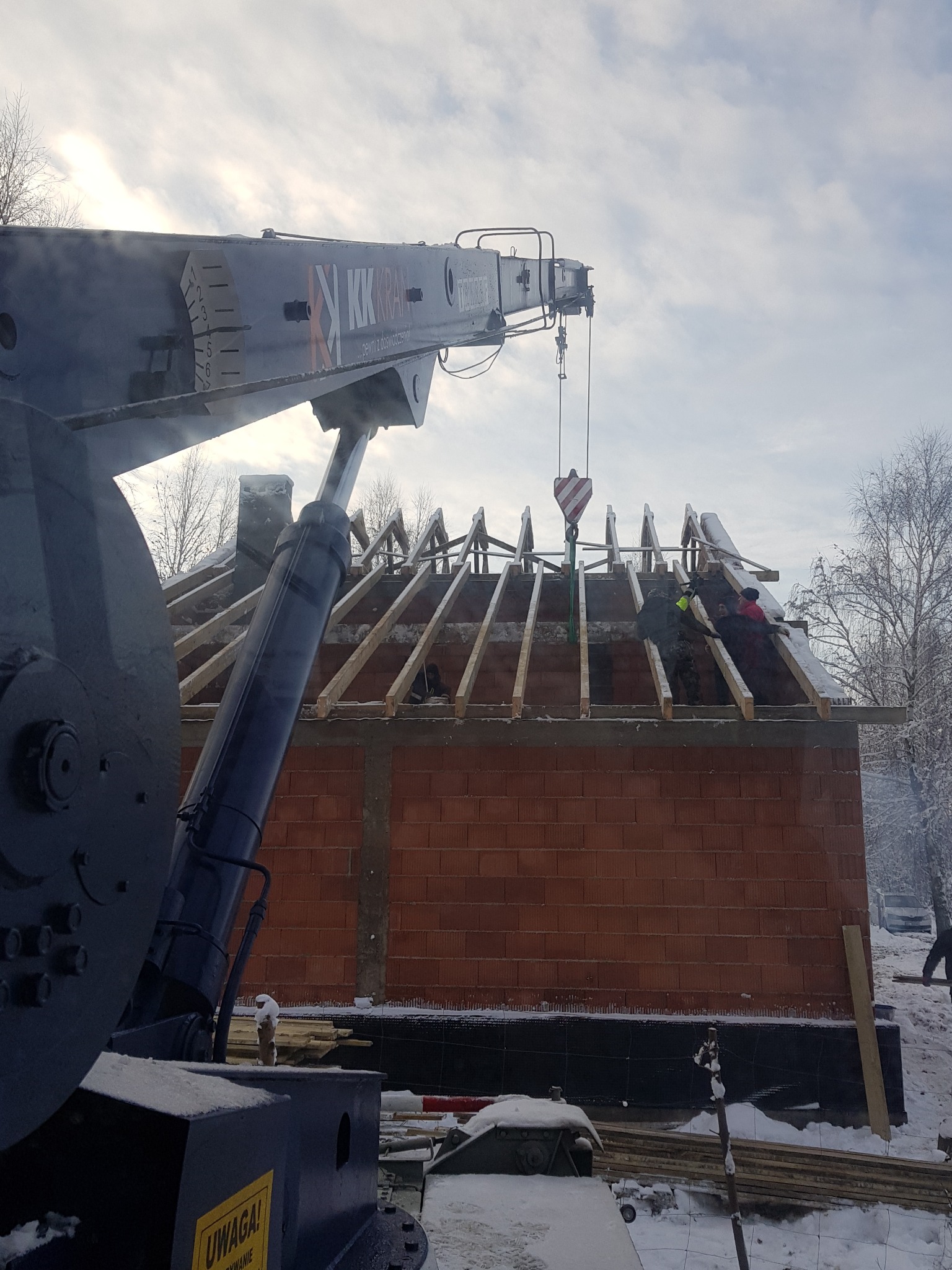 Ciężki dźwig z wysięgnikiem podnosi elementy konstrukcji dachu na budynek z czerwonej cegły w zimowej scenerii.
