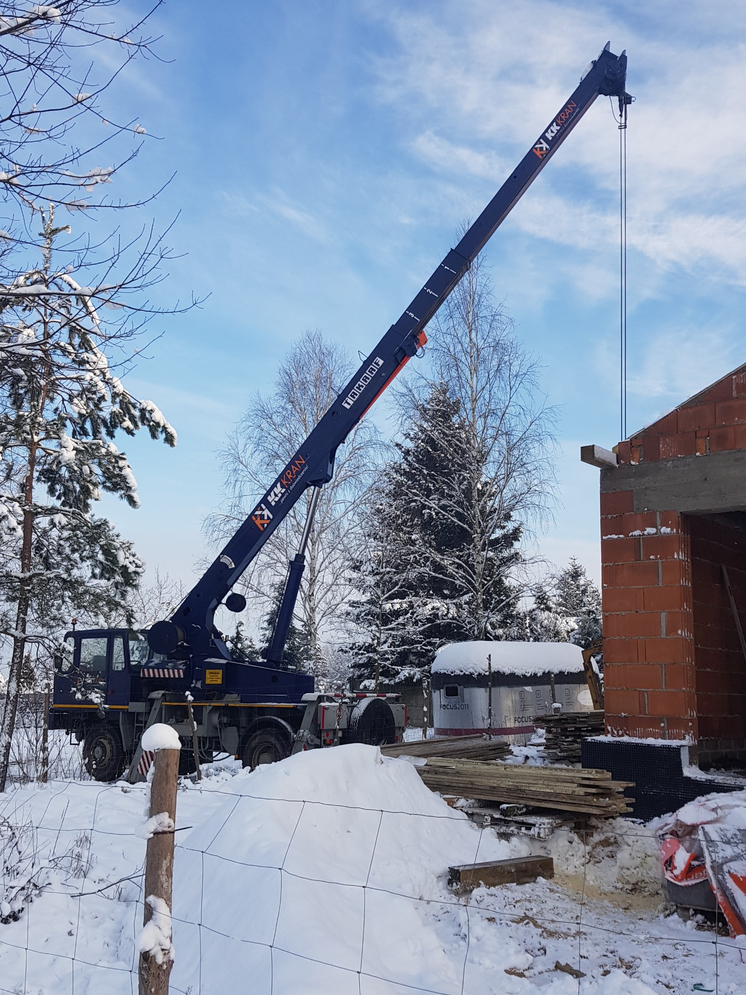 Mobilny dźwig teleskopowy z wysięgnikiem podczas podnoszenia elementu konstrukcyjnego na budowie domu jednorodzinnego w zimowej scenerii.