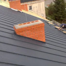 Montaż ciemnoszarego dachu panelowego z widocznym kominem z cegły i dwoma pracownikami na dachu, w tle budynki i drzewa.