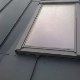 Szary dach z blachy na rąbek stojący z oknem dachowym, widoczne krople wody.